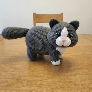Può includere: Un gatto all'uncinetto fatto a mano, grigio con accenti bianchi e rosa. Il gatto ha naso, petto e zampe bianche. Gli occhi del gatto sono neri e ha una lunga coda. Il gatto è su un tavolo di legno.