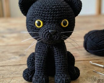 May include: A handmade black crochet cat with yellow eyes and white whiskers. The cat is sitting upright with its paws in front. A ball of black yarn and a crochet hook are in the background. This cat is a unique handmade item.