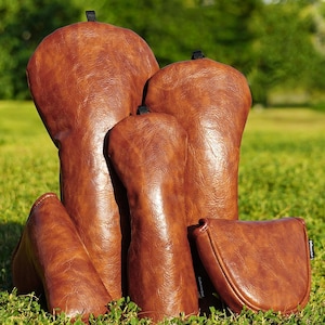 Könnte beinhalten: Eine Ansammlung brauner Golfschlägerhauben und eine Putterhaube liegen auf grünem Gras. Die Hauben bestehen aus einem strukturierten, lederartigen Material. Der Hintergrund zeigt verschwommenes grünes Laub und einen blauen Himmel.