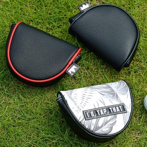 May include: Three golf putter head covers on green grass. Two are black with a crown tag, one with red trim. The third has a black and white tropical leaf pattern and the text "I'D TAP THAT". A white golf ball is also present.