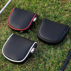 May include: Three black golf putter covers with textured surfaces are displayed on green grass. One cover has red trim, another has white trim, and the third has no trim. Each cover has a small tag with a white crown logo.