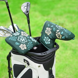 Puede incluir: Una bolsa de golf con palos y dos fundas de putter verdes. Las fundas presentan un diseño floral con flores blancas, azul claro y doradas. La bolsa de golf es blanca y negra, sobre un fondo verde.