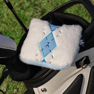 Könnte beinhalten: Eine Golftasche in Weiß und Schwarz, die Golfschläger und eine weiße, flauschige Golfschlägerhaube mit blauem Rautenmuster enthält. Die Golfschläger haben silberne und schwarze Details, vor einem grünen Gras Hintergrund.