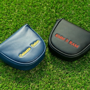 May include: Two golf club head covers on green grass. One is navy blue with gold text that reads "Celeste Eaton." The other is black with red text that reads "KMM X RAM."