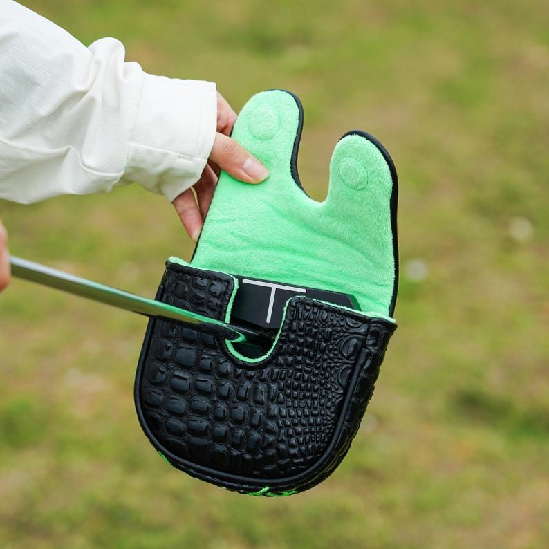 Puede incluir: Una funda negra para la cabeza del putter de golf con textura de piel de cocodrilo. El interior es de color verde brillante y la funda se mantiene abierta para revelar el putter. El fondo es hierba verde borrosa.