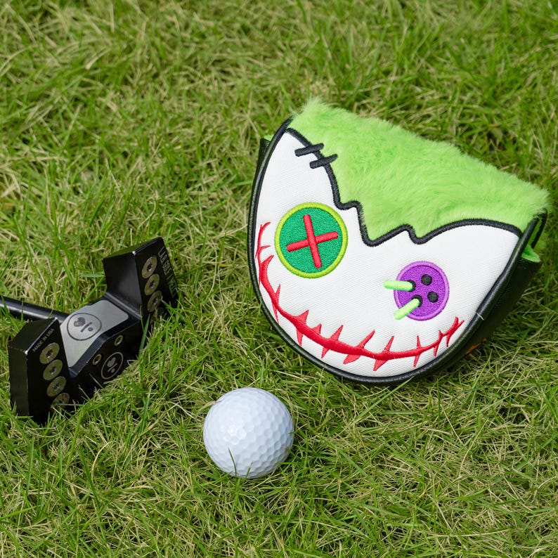 Pode incluir: Um putter de golfe preto e uma bola de golfe branca repousam sobre a relva verde ao lado de uma capa para a cabe&ccedil;a do taco de golfe branca e verde. A capa tem um design de rosto costurado com detalhes vermelhos, verdes e roxos.