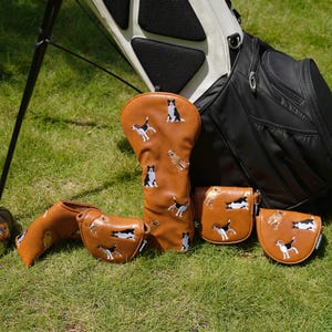 Puede incluir: Un conjunto de fundas y bolsas para palos de golf marrones, bordadas con dise&ntilde;os de perros. Los accesorios est&aacute;n colocados sobre hierba verde, junto a una bolsa de golf negra y blanca.