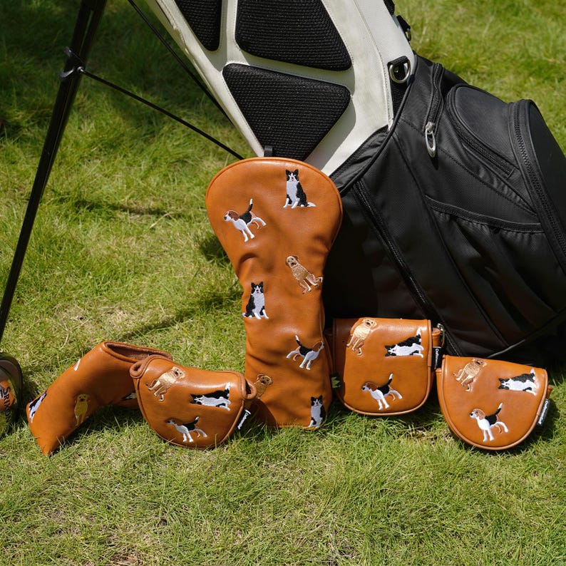 Puede incluir: Un conjunto de fundas y bolsas para palos de golf marrones, bordadas con dise&ntilde;os de perros. Los accesorios est&aacute;n colocados sobre hierba verde, junto a una bolsa de golf negra y blanca.