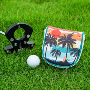 May include: A black golf putter, a white golf ball, and a golf club head cover with a tropical sunset design featuring palm trees, a surfboard, and a red sun. The putter has the text "LAB GOLF" and "DF3".