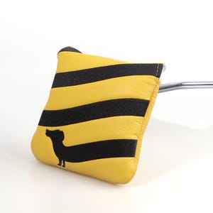 May include: Yellow and black striped golf club head cover with a black dachshund silhouette. The cover is made of a soft material and is designed to protect the club head. The cover is yellow with black stripes.