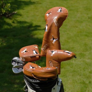 Puede incluir: Un juego de fundas para palos de golf marrones con dise&ntilde;os de perros bordados. Las fundas est&aacute;n hechas de un material similar al cuero y presentan varias razas de perros en blanco, negro y marr&oacute;n. Los palos de golf est&aacute;n en una bolsa de golf en blanco y negro.