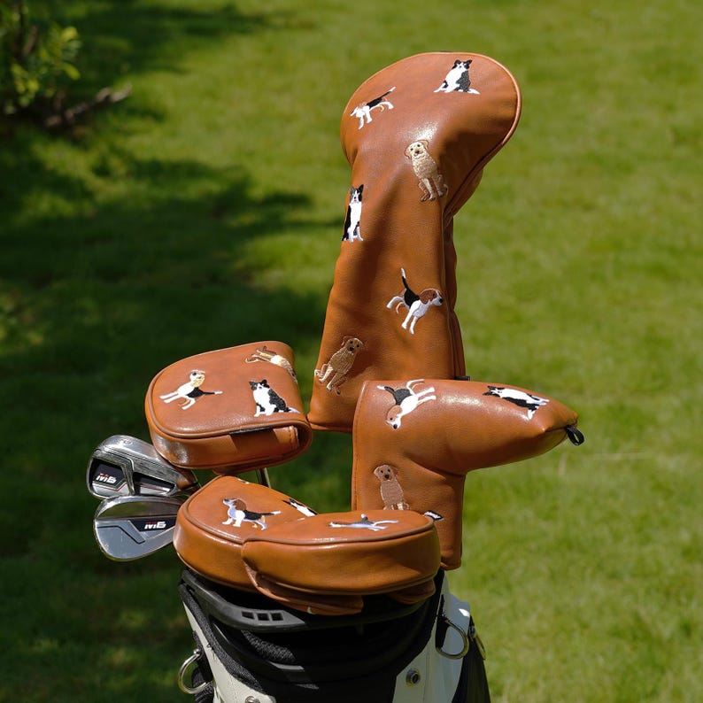 Puede incluir: Un juego de fundas para palos de golf marrones con dise&ntilde;os de perros bordados. Las fundas est&aacute;n hechas de un material similar al cuero y presentan varias razas de perros en blanco, negro y marr&oacute;n. Los palos de golf est&aacute;n en una bolsa de golf en blanco y negro.