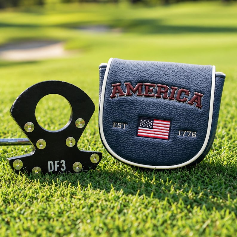 May include: A black metal putter head with the text DF3 and a navy blue textured putter cover featuring the word AMERICA in red, a small embroidered American flag, and the text EST 1776 with white piping along the curved edges of the cover.