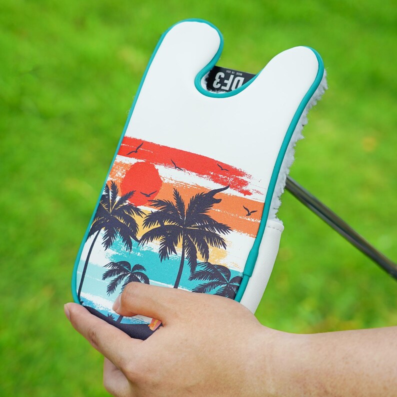 May include: White golf club head cover with a tropical sunset design featuring palm trees, a red sun, and birds. The cover has a teal trim and the text "DF3".