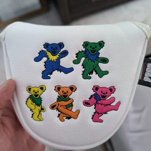 May include: White golf club head cover featuring embroidered dancing bears in blue, green, yellow, orange, and pink. Each bear has a unique pose and a colorful collar. The cover is held by a hand.