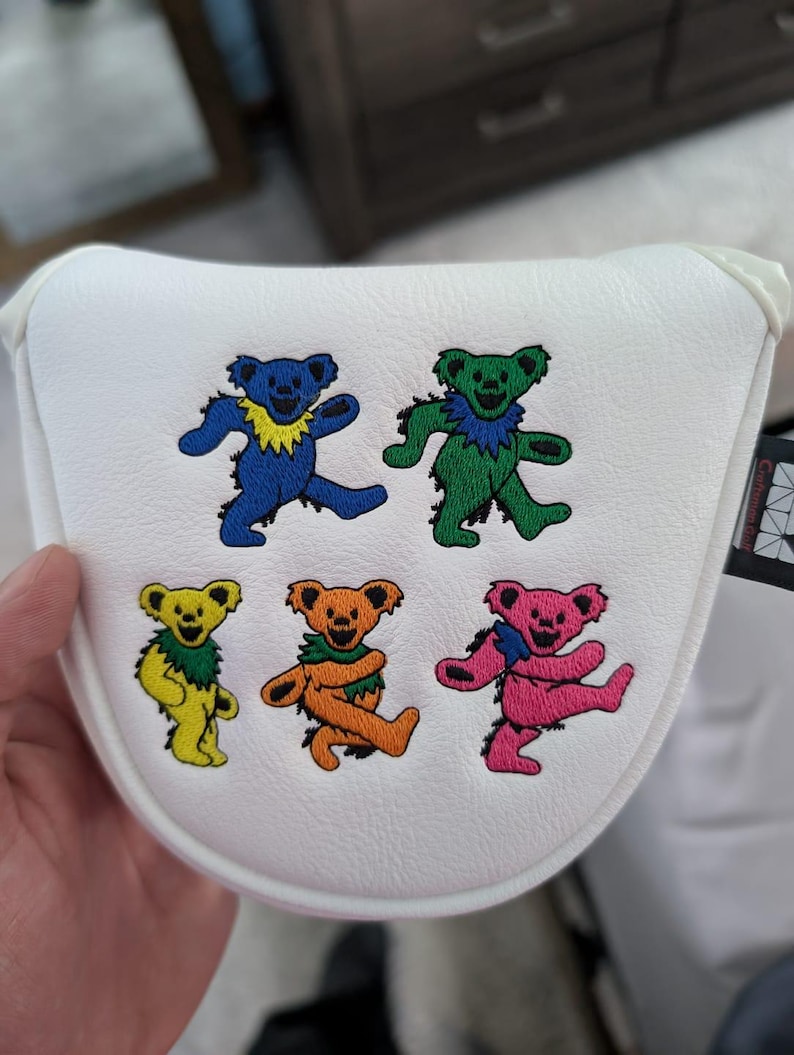 May include: White golf club head cover featuring embroidered dancing bears in blue, green, yellow, orange, and pink. Each bear has a unique pose and a colorful collar. The cover is held by a hand.