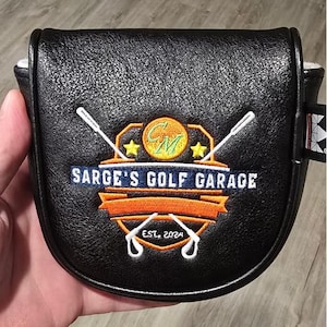 May include: Black golf putter cover with the text "SARGE'S GOLF GARAGE" and the year "EST. 2024". The cover features a logo with crossed golf clubs, stars, and a golf ball design. The cover is held in a hand.