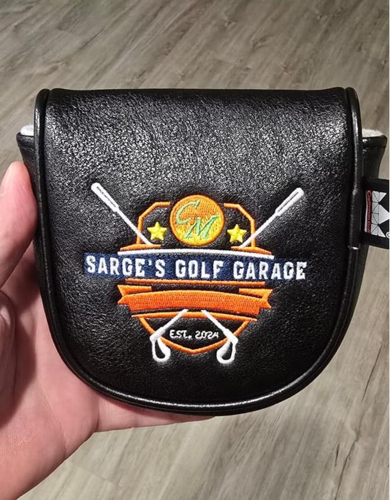 May include: Black golf putter cover with the text "SARGE'S GOLF GARAGE" and the year "EST. 2024". The cover features a logo with crossed golf clubs, stars, and a golf ball design. The cover is held in a hand.