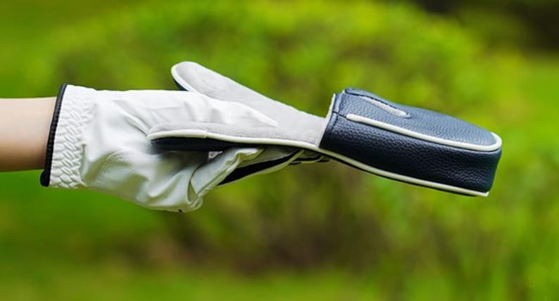 May include: A white golf glove with a black trim at the wrist and a dark blue textured golf club head cover with white piping. The head cover is held horizontally, showing its smooth interior lining and the exterior pebbled leather texture.