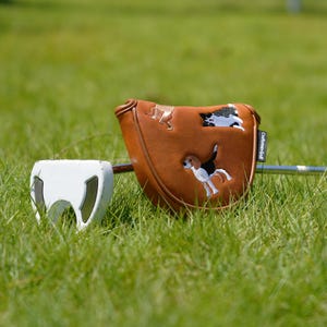 Puede incluir: Una funda de putter de golf de cuero marr&oacute;n con dise&ntilde;os de perros bordados. La funda tiene un cierre magn&eacute;tico y el texto "Craftsman Golf". Un putter de golf blanco est&aacute; al lado de la funda, sobre hierba verde.
