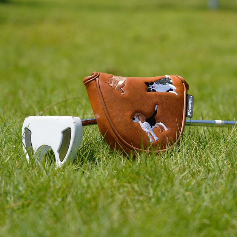 Puede incluir: Una funda de putter de golf de cuero marr&oacute;n con dise&ntilde;os de perros bordados. La funda tiene un cierre magn&eacute;tico y el texto "Craftsman Golf". Un putter de golf blanco est&aacute; al lado de la funda, sobre hierba verde.