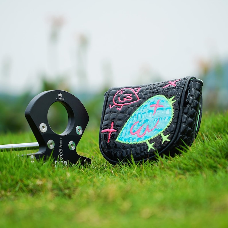 Puede incluir: Putter de golf negro con una funda a juego. El putter tiene un dise&ntilde;o circular. La funda es negra con un patr&oacute;n texturizado y presenta un dise&ntilde;o bordado colorido de un p&aacute;jaro y la palabra "Cal".