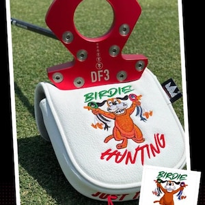 May include: A red golf putter with a white head cover featuring a cartoon dog with the words "Birdie Hunting". The putter has the text "DF3" and "LAB & GOLF" on it. The putter is on a green golf course.