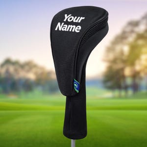 Könnte beinhalten: Schwarze Golfschlägerhaube mit dem Text "Your Name" in Weiß. Die Hülle hat ein Mesh-Panel und eine gerippte Stricksocke. Ein kleines grünes und blaues Logo befindet sich an der Seite. Der Hintergrund ist ein Golfplatz.