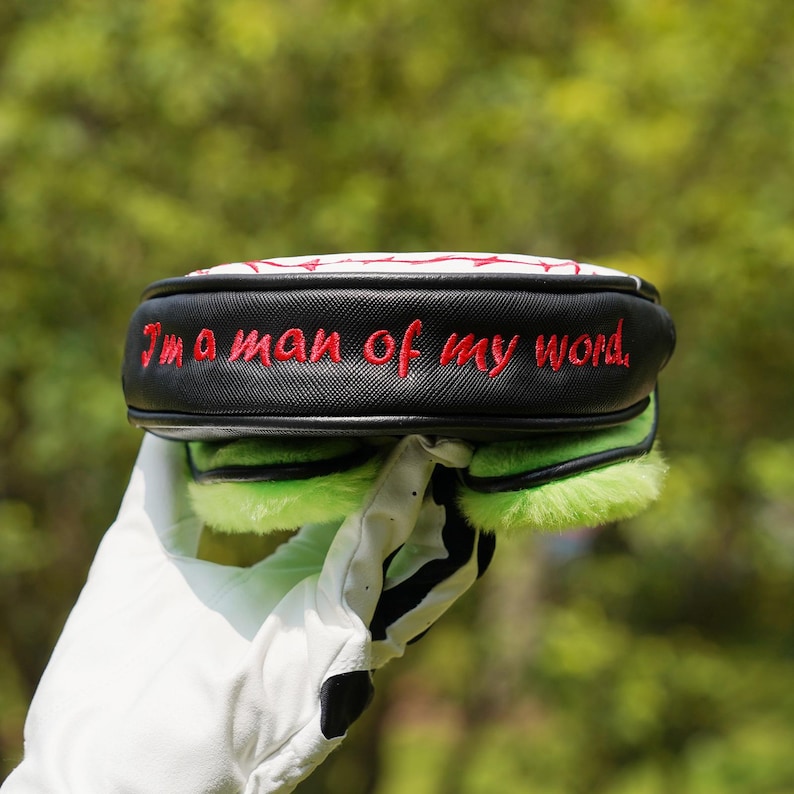 Pode incluir: Uma capa para a cabe&ccedil;a do taco de golfe em preto e branco com a frase bordada "I'm a man of my word" em vermelho. A capa tem um topo branco com costuras vermelhas e detalhes difusos verdes.