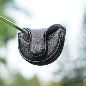 Może przedstawiać: Czarna sk&oacute;rzana osłona gł&oacute;wki puttera golfowego o teksturowanej powierzchni. Osłona ma chronić gł&oacute;wkę puttera i ma zakrzywiony kształt. Widoczny jest trzonek puttera.