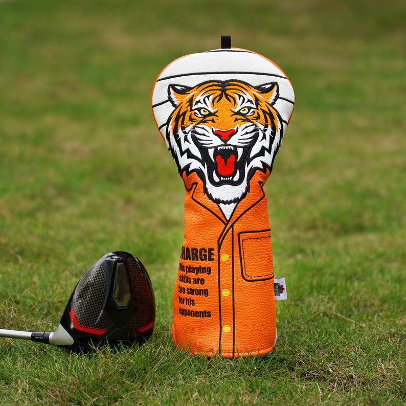 Op de afbeelding: Een oranje en witte golfclubhoofdbeschermer met een tijgerontwerp. De hoes heeft een tijgergezicht aan de bovenkant en de tekst "MARGE" met een citaat over speelvaardigheden aan de voorkant. Een zwarte en rode golfclub ligt in het gras.