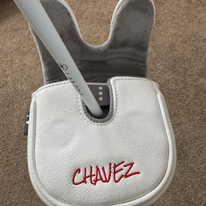 May include: A white golf putter head cover with the name "CHAVEZ" embroidered in red. The cover is made of a textured material and has a grey, plush interior. A white golf club is inserted into the cover.