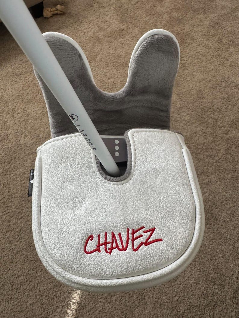 May include: A white golf putter head cover with the name "CHAVEZ" embroidered in red. The cover is made of a textured material and has a grey, plush interior. A white golf club is inserted into the cover.