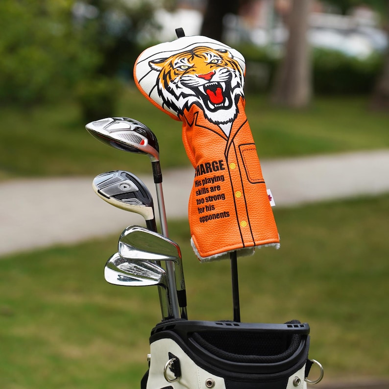 Op de afbeelding: Golfclub hoes met een tijgerontwerp. De oranje hoes heeft een tijgergezichtafbeelding en de tekst "MARGE His playing skills are too strong for his opponents". De golfclubs zitten in een zwart-witte golftas.