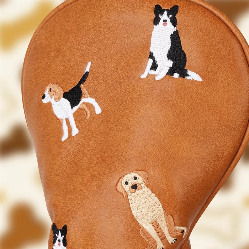 Puede incluir: Una funda marr&oacute;n para la cabeza del palo de golf con bordados de perros. La funda muestra un beagle, un border collie y un golden retriever, entre otras razas de perros. La funda tiene una textura suave.