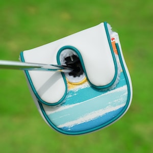 May include: White golf club head cover with a teal trim and a painted blue and white striped design. The cover has a soft, fluffy interior and a teal outline. A golf club is inserted into the cover.