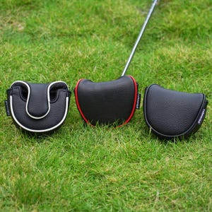 Może przedstawiać: Trzy osłony głowicy puttera golfowego na zielonej trawie. Jedna jest czarna z białym wykończeniem, druga czarna z czerwonym wykończeniem, a trzecia jednolita czarna. Pokrowce mają teksturowany wygląd i widoczna jest nazwa marki "Craftsman Golf".