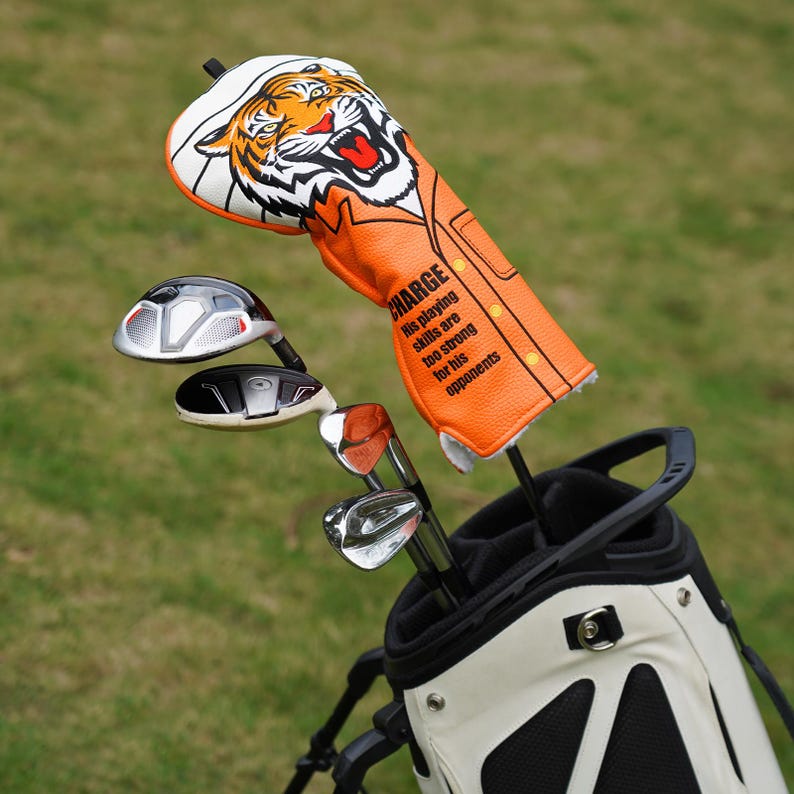 Op de afbeelding: Een golfclubset in een witte en zwarte golftas. De set bevat een driver met een tijgerhoofdbeschermer en de tekst "CHARGE". De tijgerhoofdbeschermer is oranje en wit.