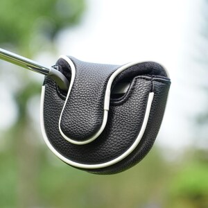 Może przedstawiać: Czarna osłona gł&oacute;wki puttera golfowego z białym wykończeniem. Osłona wykonana jest z teksturowanego materiału i ma chronić gł&oacute;wkę puttera. Putter jest częściowo włożony w osłonę, prezentując jej design i funkcjonalność. Tło jest rozmyte.