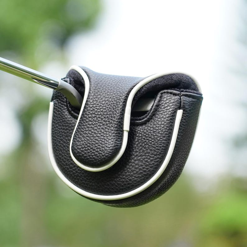 Może przedstawiać: Czarna osłona gł&oacute;wki puttera golfowego z białym wykończeniem. Osłona wykonana jest z teksturowanego materiału i ma chronić gł&oacute;wkę puttera. Putter jest częściowo włożony w osłonę, prezentując jej design i funkcjonalność. Tło jest rozmyte.