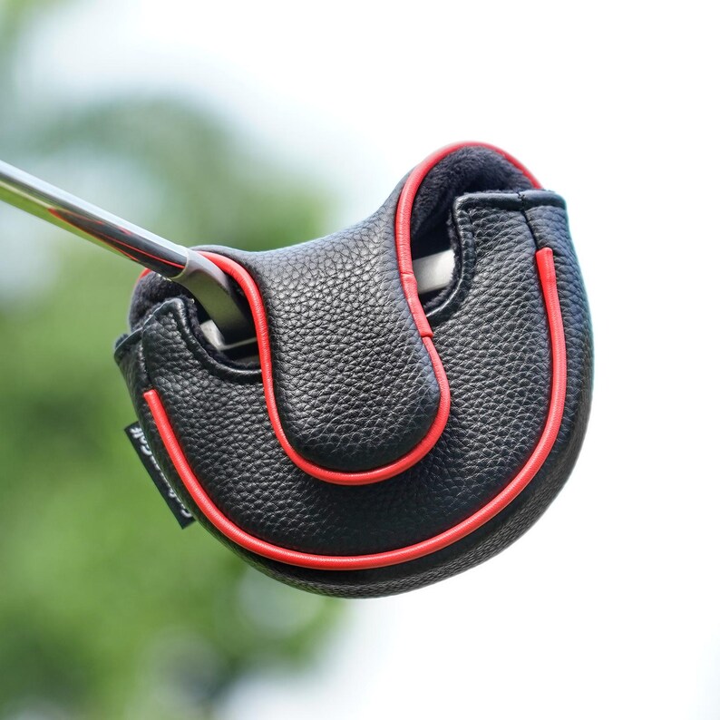 Może przedstawiać: Czarny sk&oacute;rzany pokrowiec na gł&oacute;wkę puttera golfowego z czerwonymi lam&oacute;wkami. Pokrowiec ma chronić gł&oacute;wkę puttera i ma teksturowaną powierzchnię. Putter jest włożony do pokrowca, na rozmytym zielonym tle.
