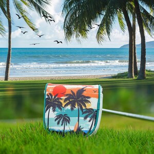 May include: A white golf bag with a colorful beach scene featuring palm trees, a red sun, and a surfboard. The bag has a teal trim and is set against a backdrop of a beach and ocean with palm trees.