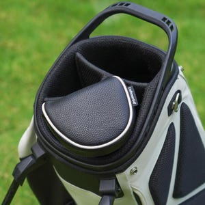 Może przedstawiać: Torba golfowa w kolorach czarnym i białym z czarnym uchwytem. Torba posiada czarną osłonę ze sk&oacute;ry ekologicznej z białym wykończeniem. Torba znajduje się na zielonym tle.