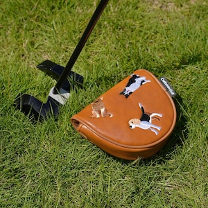 Puede incluir: Una funda para la cabeza del palo de golf de cuero marr&oacute;n con dise&ntilde;os de perros bordados. La funda est&aacute; sobre una superficie de hierba verde con un palo de golf negro en el fondo. El texto "Craftsman Golf" es visible.