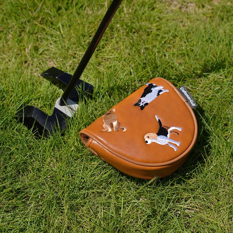 Puede incluir: Una funda para la cabeza del palo de golf de cuero marr&oacute;n con dise&ntilde;os de perros bordados. La funda est&aacute; sobre una superficie de hierba verde con un palo de golf negro en el fondo. El texto "Craftsman Golf" es visible.