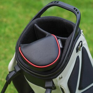 Może przedstawiać: Torba golfowa w kolorze czarnym i białym, z czarną osłoną o efekcie sk&oacute;ry z czerwonym obramowaniem. Torba posiada uchwyt i kilka przegr&oacute;dek na kije. Widoczna jest nazwa marki "Craftsman Golf".