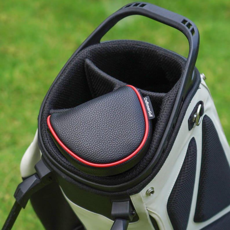 Może przedstawiać: Torba golfowa w kolorze czarnym i białym, z czarną osłoną o efekcie sk&oacute;ry z czerwonym obramowaniem. Torba posiada uchwyt i kilka przegr&oacute;dek na kije. Widoczna jest nazwa marki "Craftsman Golf".