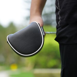 Może przedstawiać: Czarna osłona gł&oacute;wki puttera golfowego z białym wykończeniem. Osłona ma teksturowaną powierzchnię, a na boku nadrukowano napis "Craftsman Golf". Osłona jest trzymana w dłoni osoby.