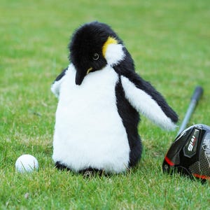 Puede incluir: Una funda para la cabeza del palo de golf con forma de pingüino en negro, blanco y amarillo, colocada sobre hierba verde junto a una pelota de golf y un palo de golf. La funda tiene un diseño realista.