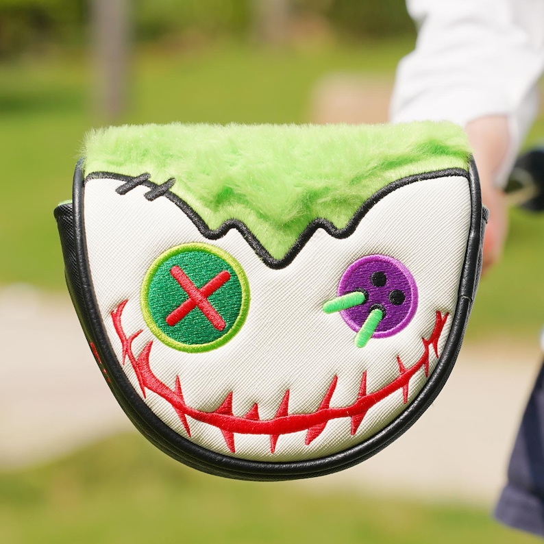 Pode incluir: Uma capa para a cabe&ccedil;a do taco de golfe com um design de rosto de monstro. A capa apresenta um topo verde e difuso, um rosto branco com detalhes costurados e uma borda preta. O rosto tem uma boca costurada vermelha e olhos de bot&atilde;o verdes e roxos.